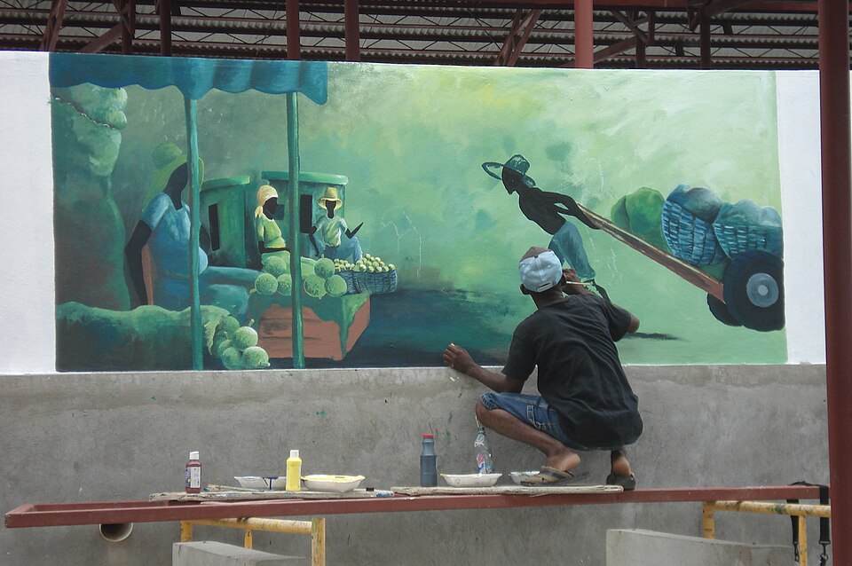 Haitian artist painting a mural of a market scene with vendors and a man pulling a cart